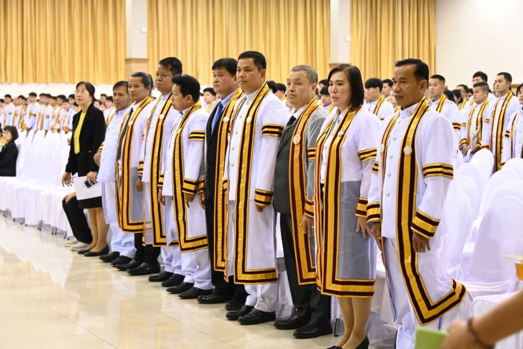 ผู้ทรงคุณวุฒิและบัณฑิตกิตติมศักดิ์ในชุดครุยสีขาว ยืนเคารพเพลงชาติหรือเพลงสรรเสริญพระบารมี บริเวณที่นั่งแถวหน้าในหอประชุมพิธีพระราชทานปริญญาบัตร