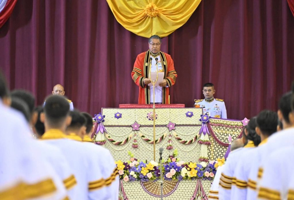 นายพลากร สุวรรณรัฐ องคมนตรี ในชุดครุยแดง กล่าวในพิธีพระราชทานปริญญาบัตรบนเวทีอย่างเป็นทางการ หน้าฉากม่านแดงและฉากประดับลวดลายดอกไม้สีม่วงทอง